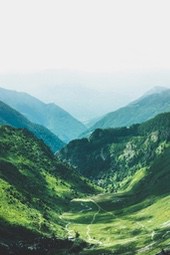 An aerial view of a valley with winding roads on green slopes, distant mountains are shrouded in mist. - free wallpaper image