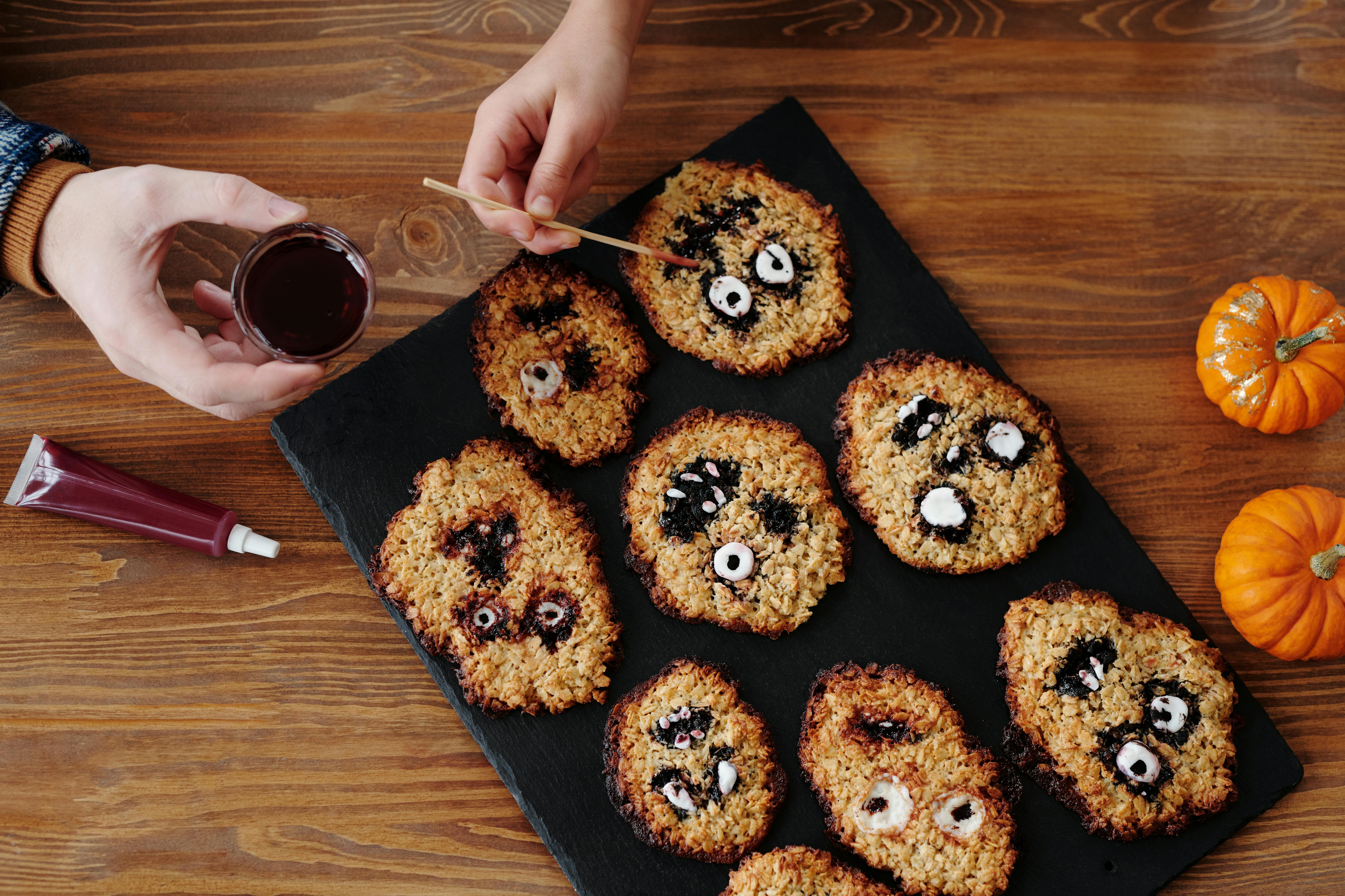 There are some ghost face cookies on a wooden table, with pumpkins and a bottle of red sauce next to them. - wallpaper image