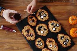 There are some ghost face cookies on a wooden table, with pumpkins and a bottle of red sauce next to them. - wallpaper image