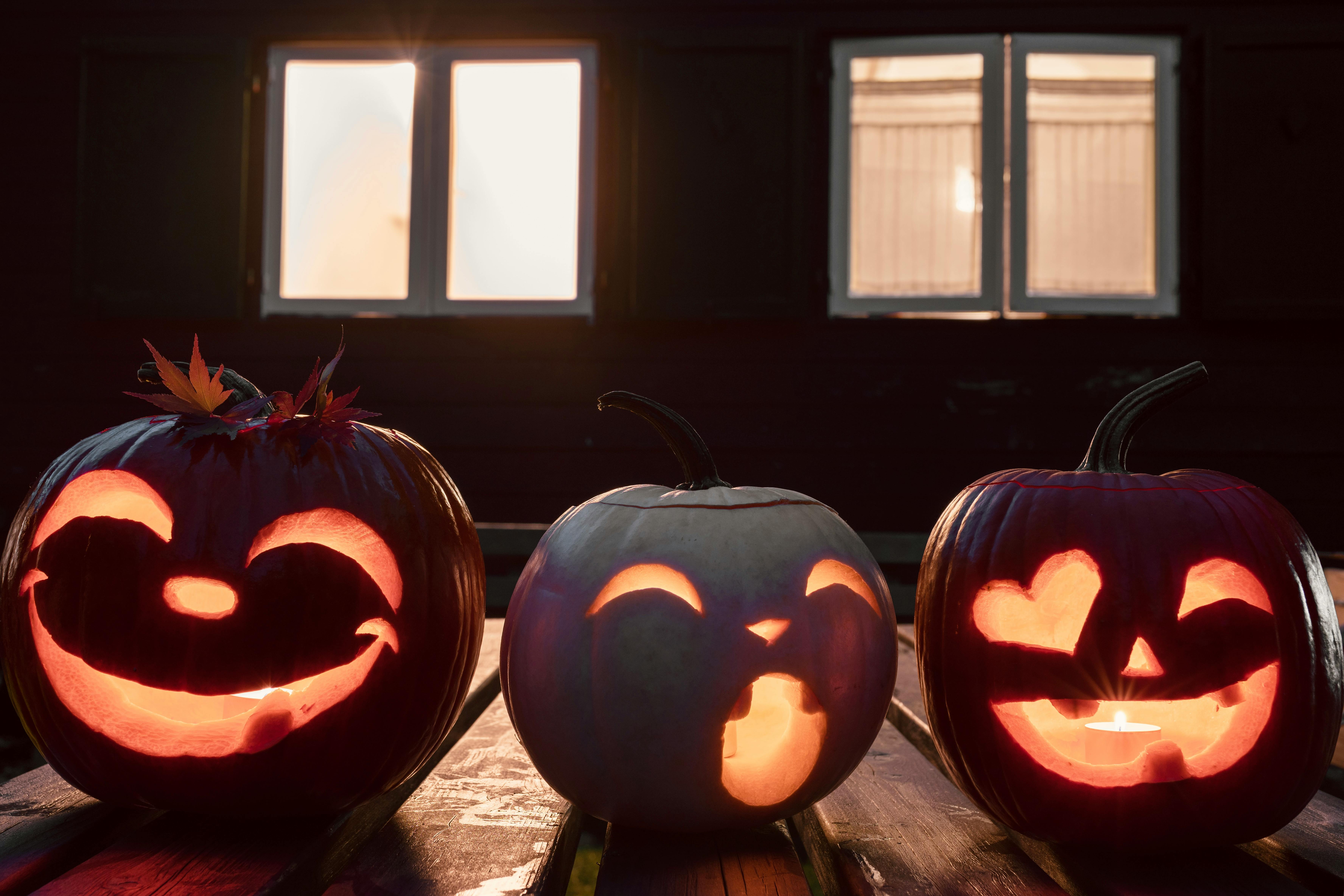 Three jack-o&apos;-lanterns are lined up on a wooden table, each with a different expression carved into them, with windows in the background and dim lighting. - wallpaper image