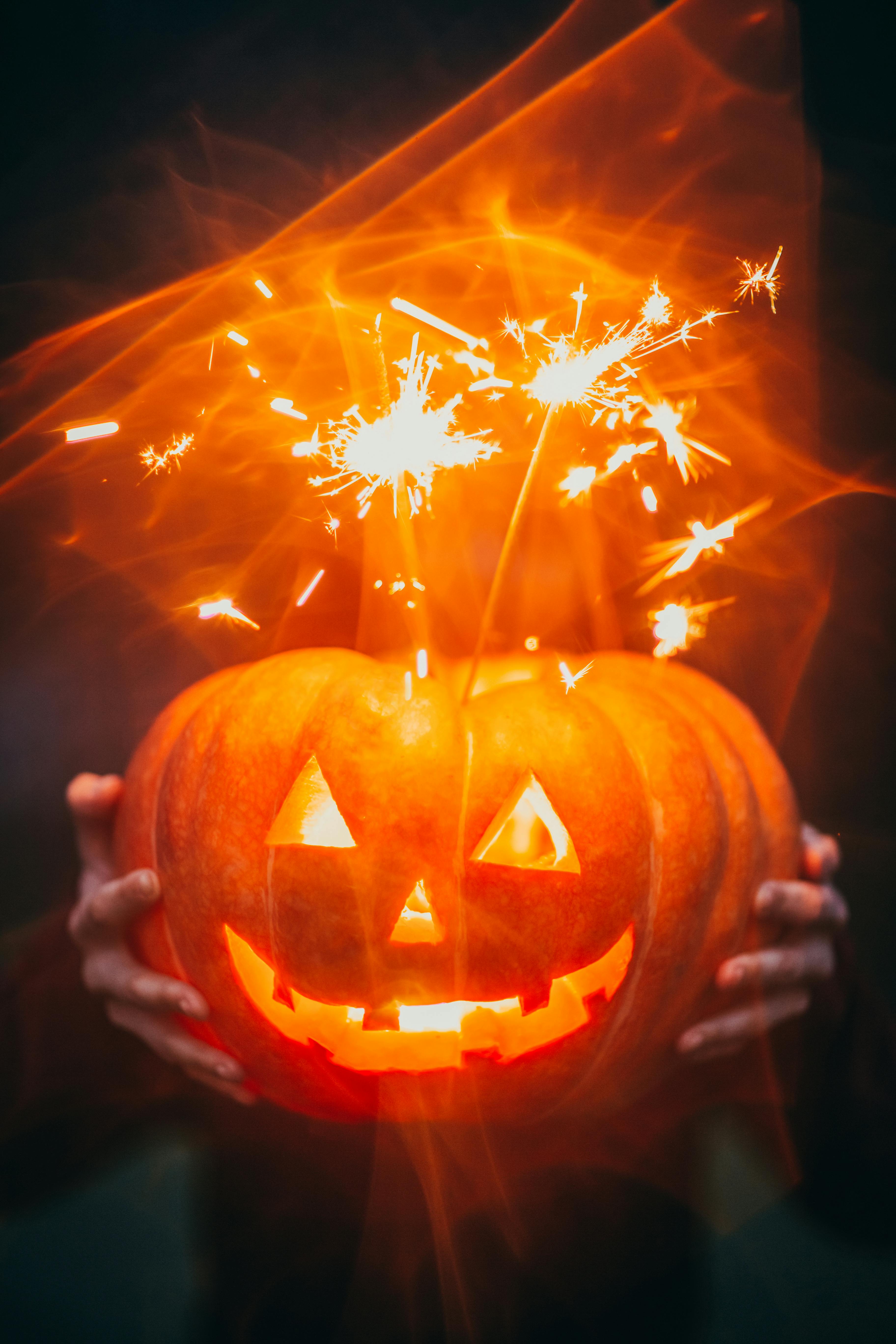 A jack-o&apos;-lantern is held up by two large hands, surrounded by red sparks. - wallpaper image