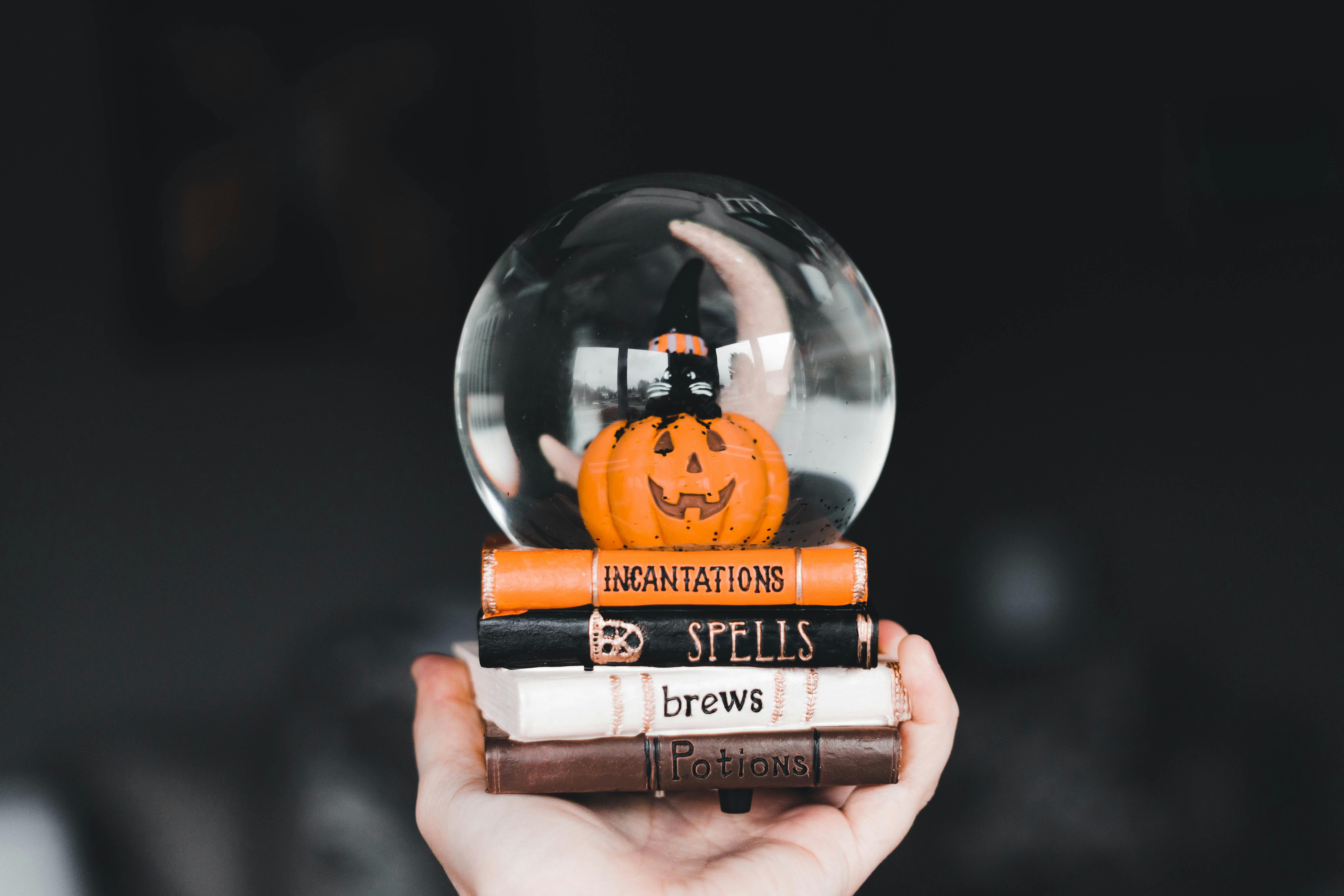 A pumpkin is placed in a glass ball, which is placed on four books with titles “Incantations”, “Spells”, “brews” and “Potions”. - wallpaper image