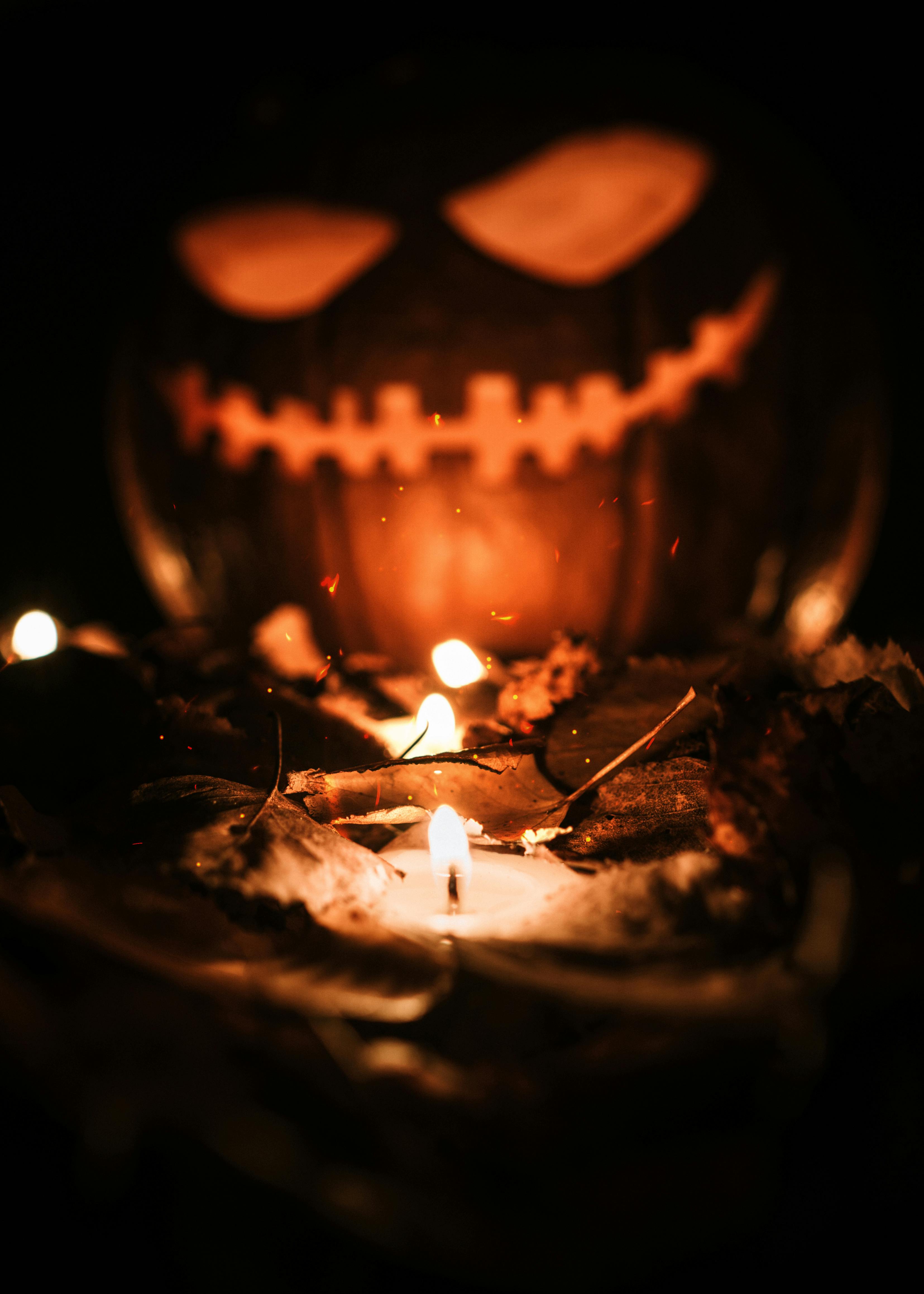 A lit pumpkin lantern emits an eerie glow in the darkness, surrounded by leaves and candles, creating a mysterious atmosphere. - wallpaper image