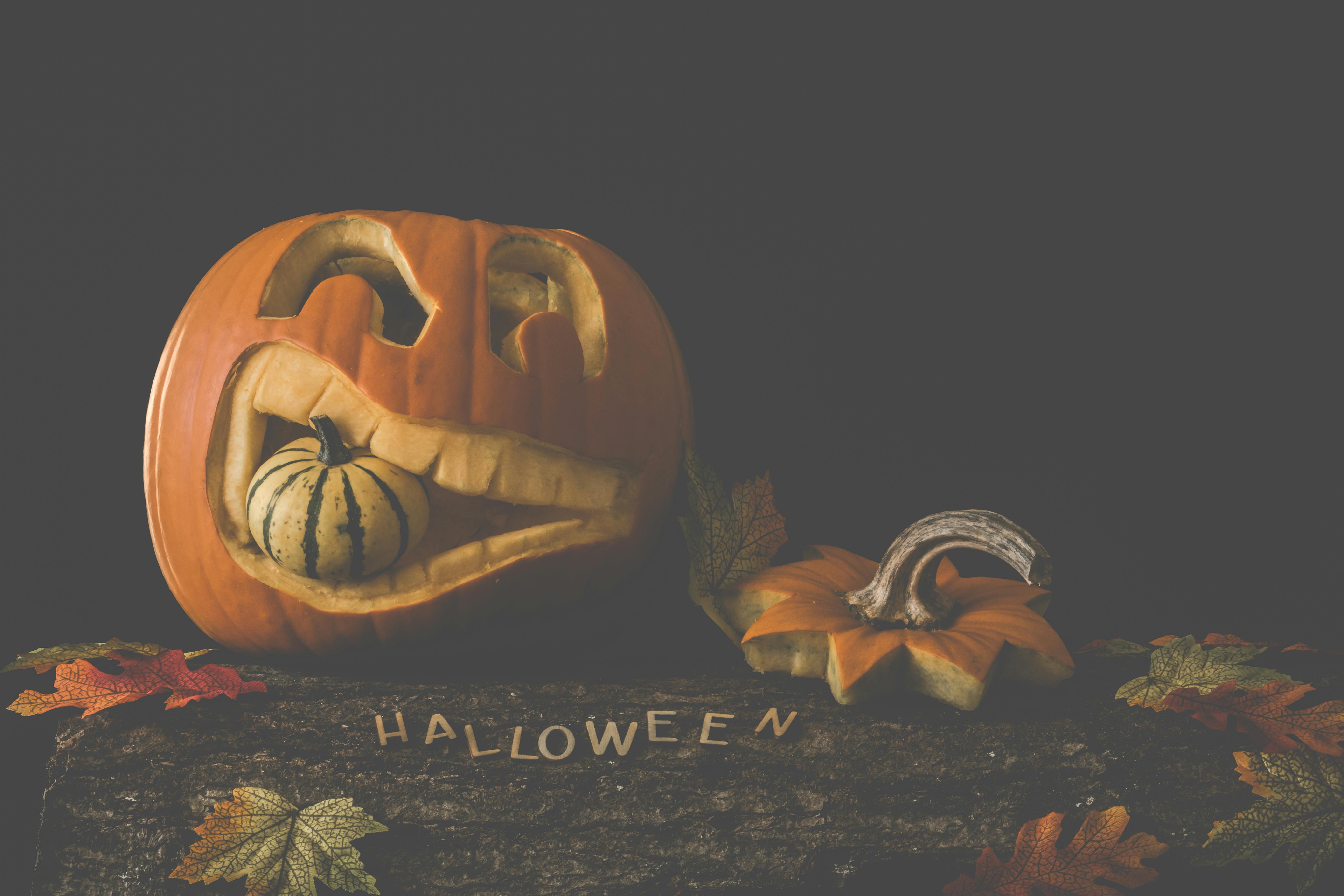 A carved pumpkin with a scary face, it has a small pumpkin in its mouth, next to it is the top of the pumpkin, below it is a wooden background and some fallen leaves, with the word &apos;Halloween&apos; written on it. - wallpaper image