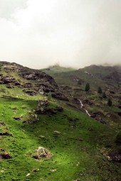 The hillside is covered with green grass, the distant peaks are shrouded in fog, and a small stream winds its way down the hillside. - free wallpaper image