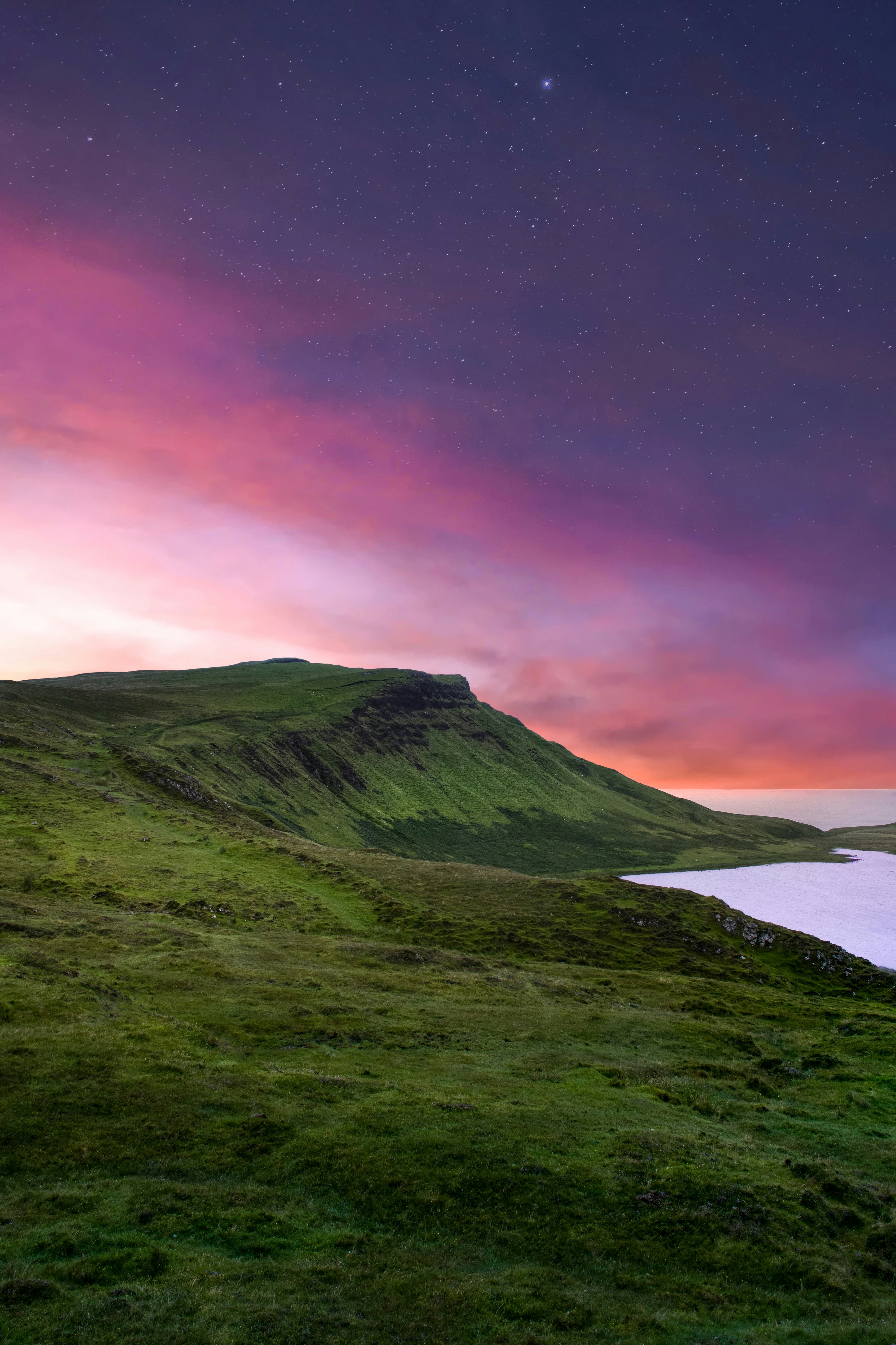 A grassy hillside with a calm lake in the distance, the sky is full of stars and pink clouds, the scene is tranquil and beautiful. - free wallpaper image