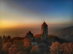 A castle stands on a hilltop at sunset, surrounded by dense forest, with a panoramic view of the city in the distance. - free wallpaper image