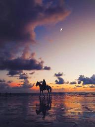 A person riding a horse on the beach at sunset, with clouds and moon in the sky, and the sea in the distance. - free wallpaper image