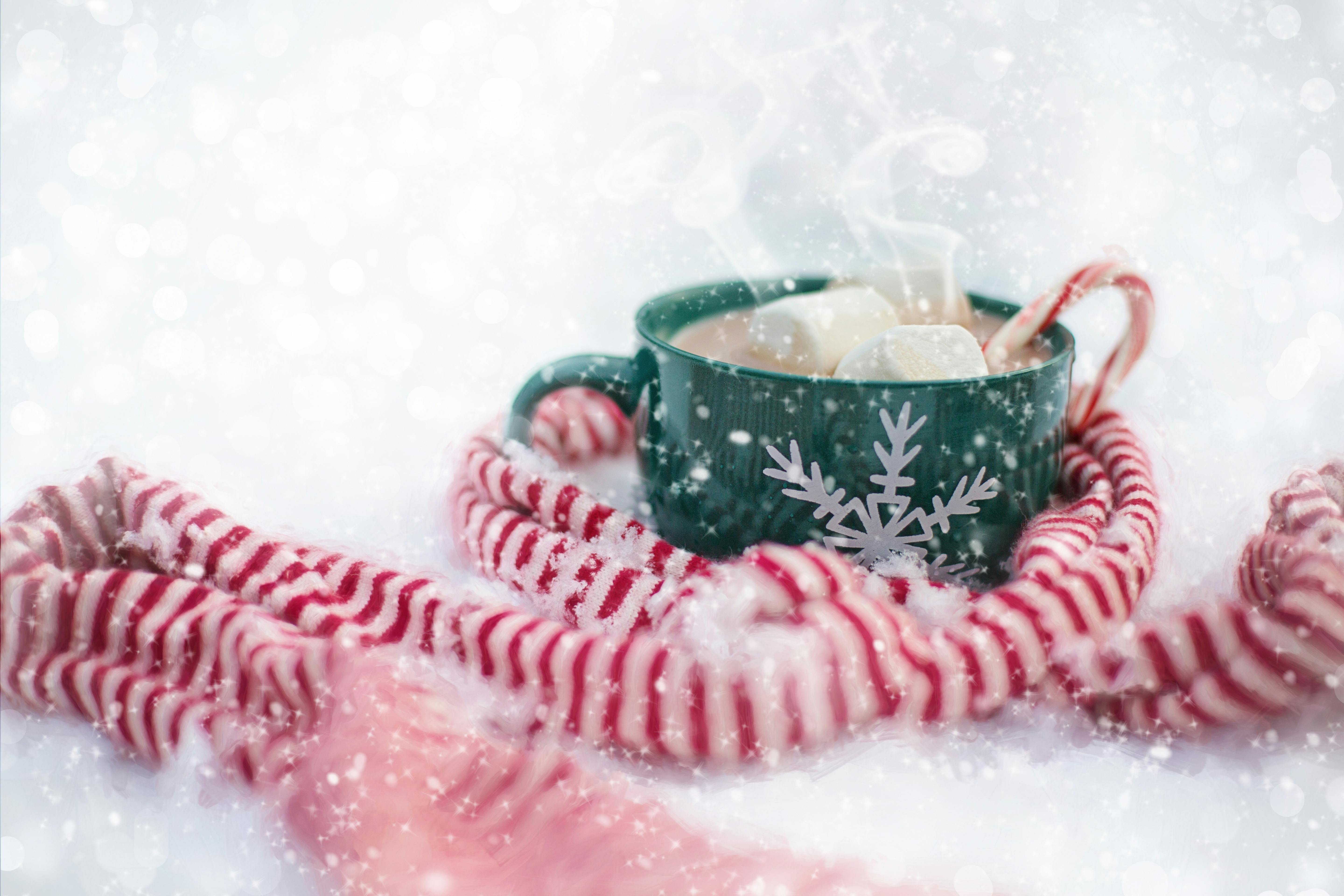 A steaming cup of hot chocolate topped with marshmallows and a candy cane, with a red and white scarf beside it. The whole scene is covered in snowflakes, creating a warm and cozy winter atmosphere. - wallpaper image