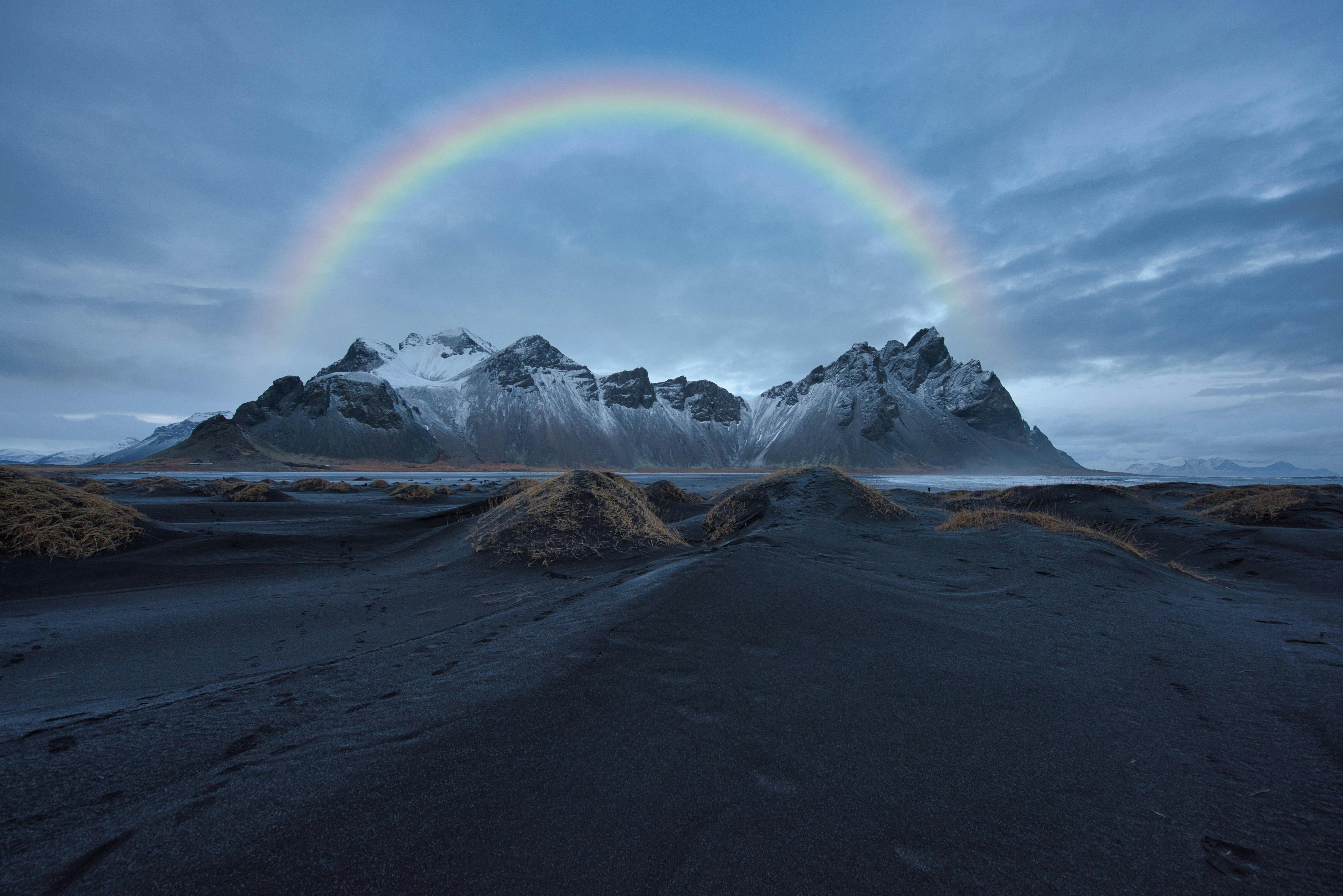 A rainbow arches over a snowy mountain range in the distance, with a black sand beach in the foreground. - free wallpaper image