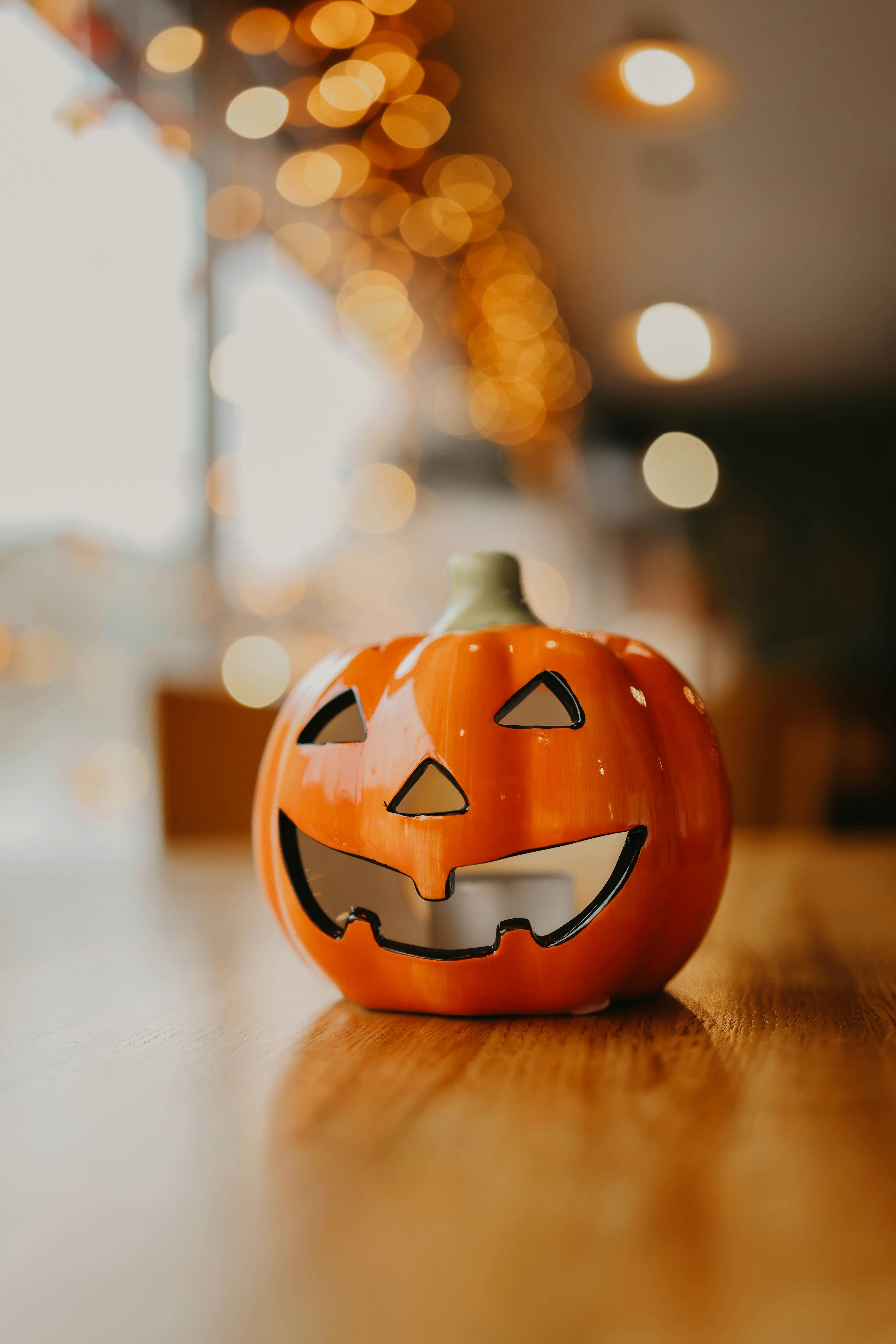 An orange jack-o&apos;-lantern sits on a wooden table with blurry lights in the background. - wallpaper image