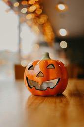 An orange jack-o&apos;-lantern sits on a wooden table with blurry lights in the background. - wallpaper image