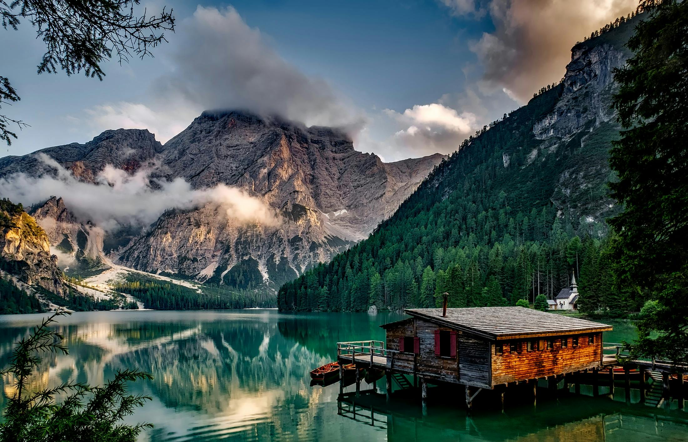 A wooden house built on the lake, surrounded by towering mountains covered in clouds, the lake water is clear and green, the scenery is picturesque. - free wallpaper image