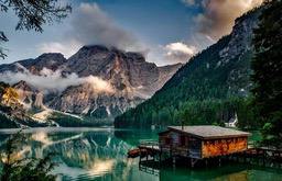 A wooden house built on the lake, surrounded by towering mountains covered in clouds, the lake water is clear and green, the scenery is picturesque. - free wallpaper image