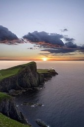 A lighthouse stands on a cliff overlooking the ocean at sunset, with clouds filling the sky. The view is majestic. - free wallpaper image