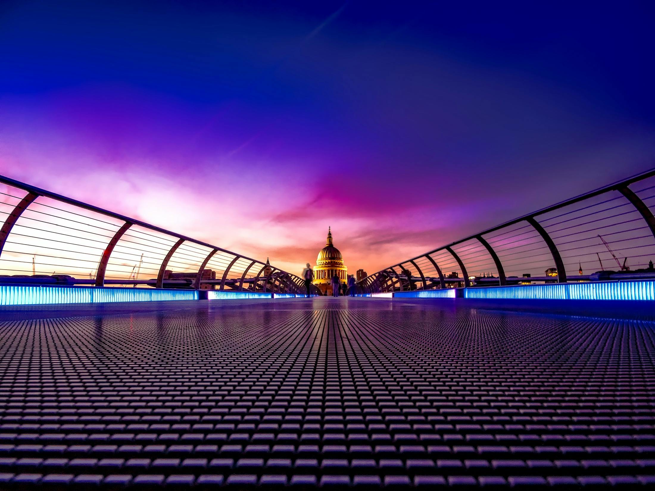 A steel bridge with a metal grid floor, in the distance is St. Paul's Cathedral in London, the sky is a gradient of orange, purple and blue. - free wallpaper image