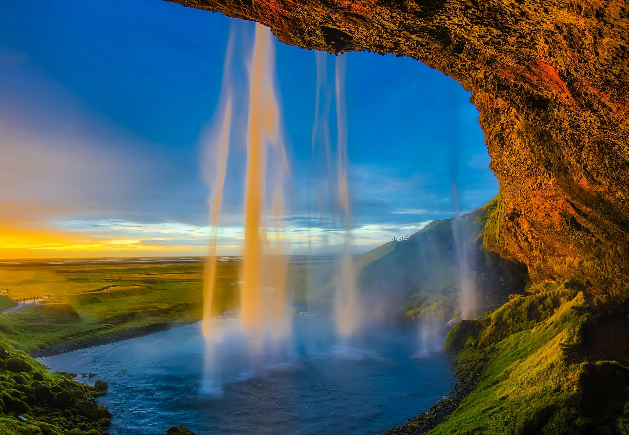 A waterfall cascades down a cliff, the sunlight shines on the water creating rainbows, the distant hillsides are covered with green vegetation, the scenery is magnificent. - free wallpaper image