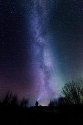 The Milky Way stretches across the night sky like a bright river, with silhouettes of trees below and a person shining a spotlight up towards the sky, with distant lights. - free wallpaper image