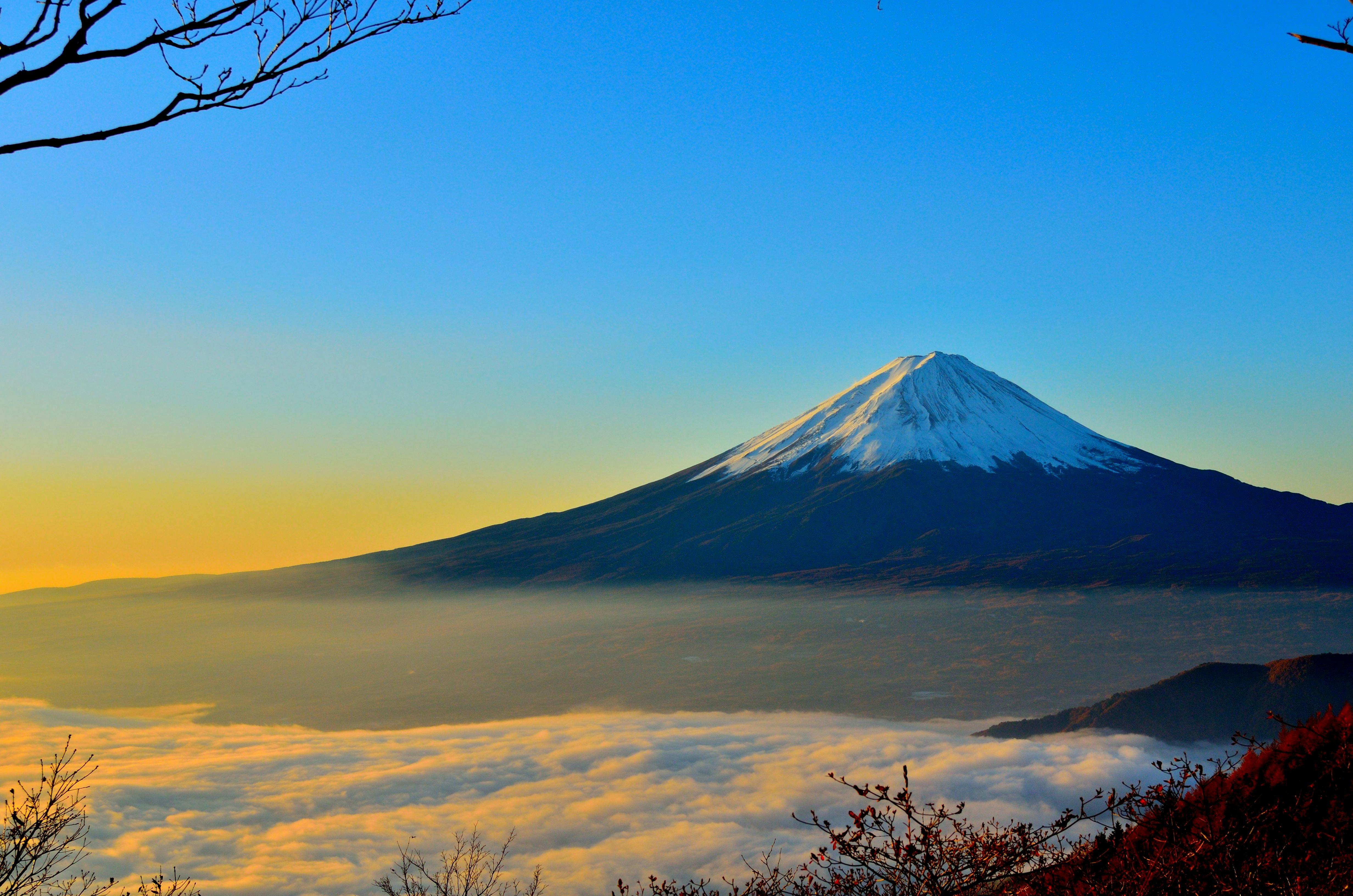 Mount Fuji is covered in snow at the top, with a sea of clouds surrounding the peak. The distant sky is a gradient of blue and yellow. - free wallpaper image