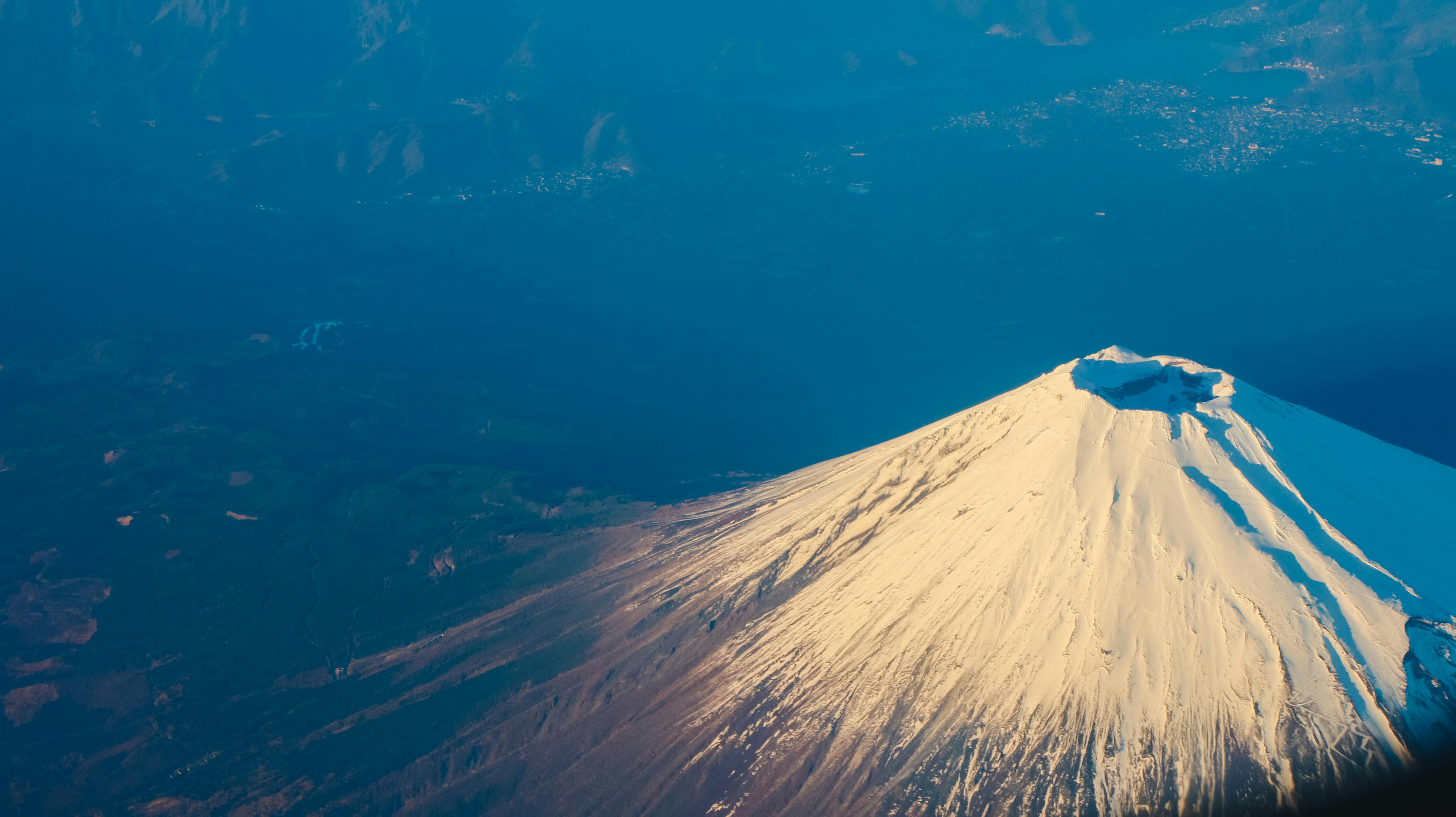 An aerial view of Mount Fuji, the summit is covered with white snow, surrounded by blue sky and green vegetation. - free wallpaper image