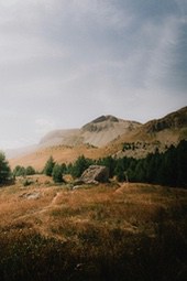 A grassy meadow in the mountains, with rolling hills in the distance. The sky is covered with clouds and sunlight shines through the clouds. - free wallpaper image