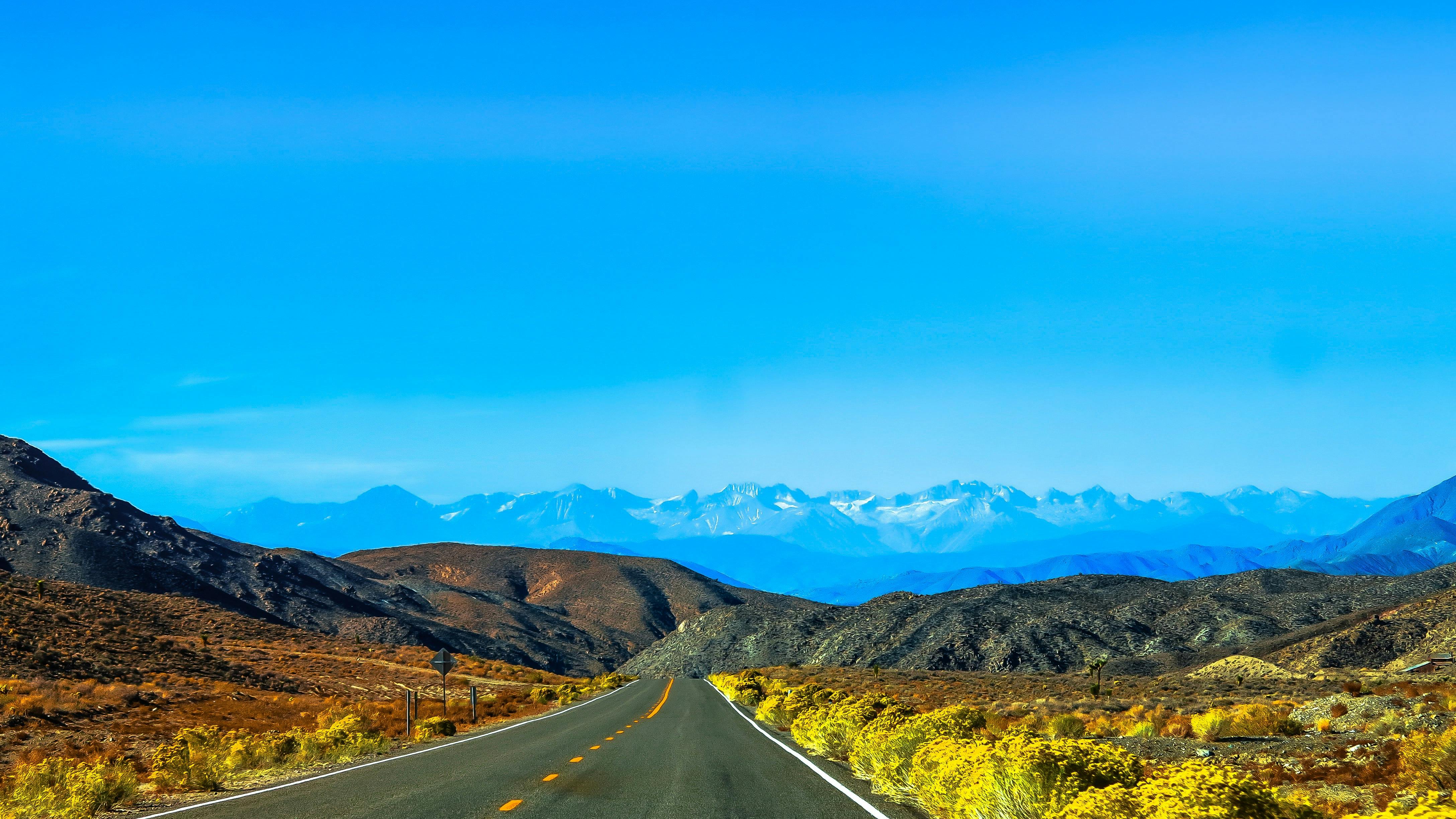 A straight road winds through the mountains, with rolling hills in the distance, a clear blue sky and a magnificent view. - free wallpaper image