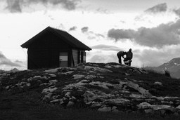 Two men stand in front of a cabin on a mountain top with mountains and sky in the background. - free wallpaper image