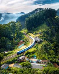 A train runs through mountainous terrain, surrounded by lush forests and tea plantations, with misty mountains in the distance. - free wallpaper image