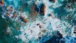 An aerial view of the sea hitting rocks, forming white waves. The sea is deep blue and the rocks are brown. - free wallpaper image