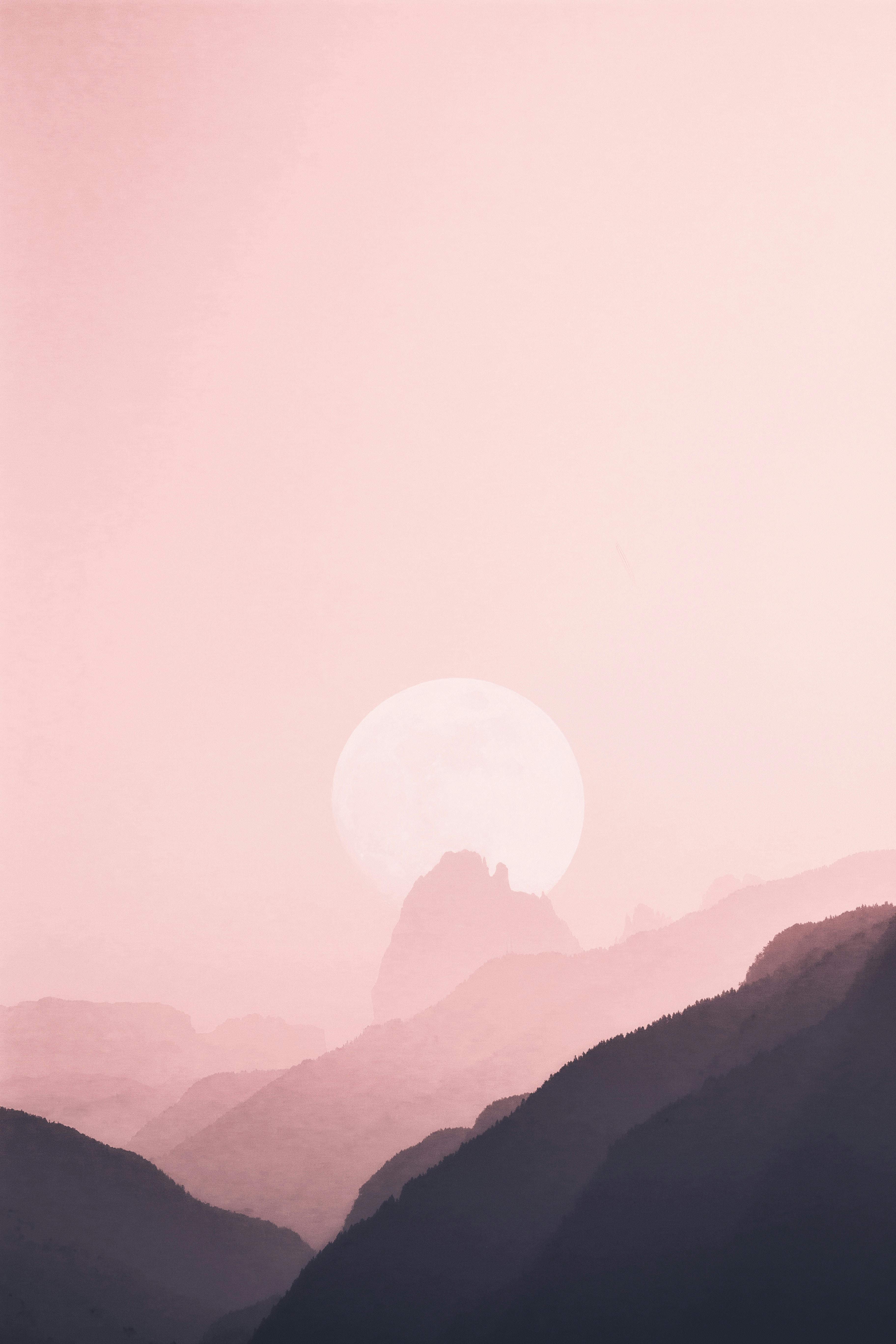 The mountains are layered under a pink sky, with a round sun/moon in the distance, it looks like sunrise/moonrise. - free wallpaper image