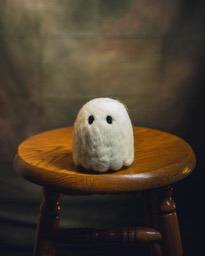 A plush ghost toy sits on a wooden stool, with a blurred wall in the background. - wallpaper image