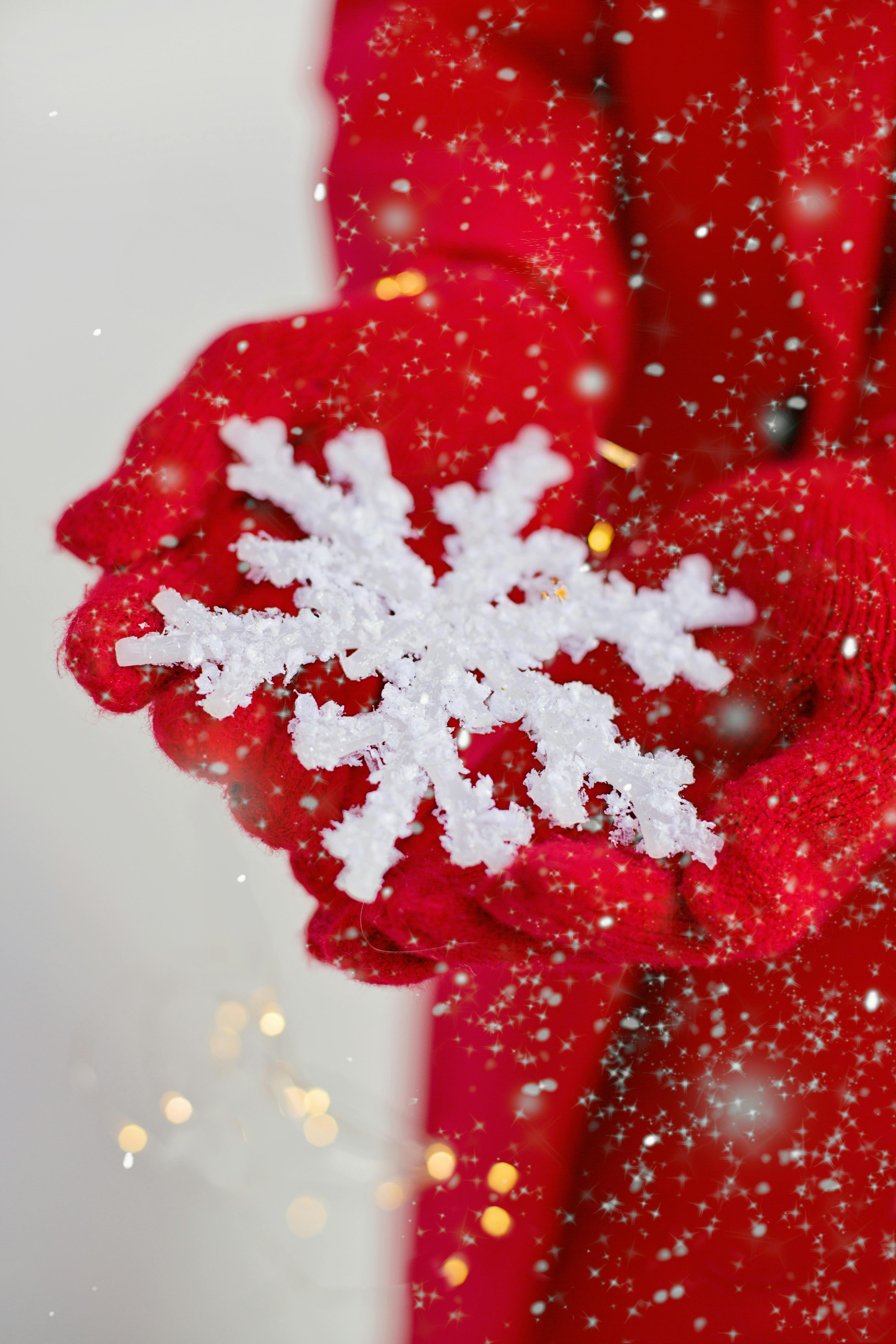 A pair of hands wearing red gloves holding a white snowflake, the background is a white snow scene. - wallpaper image