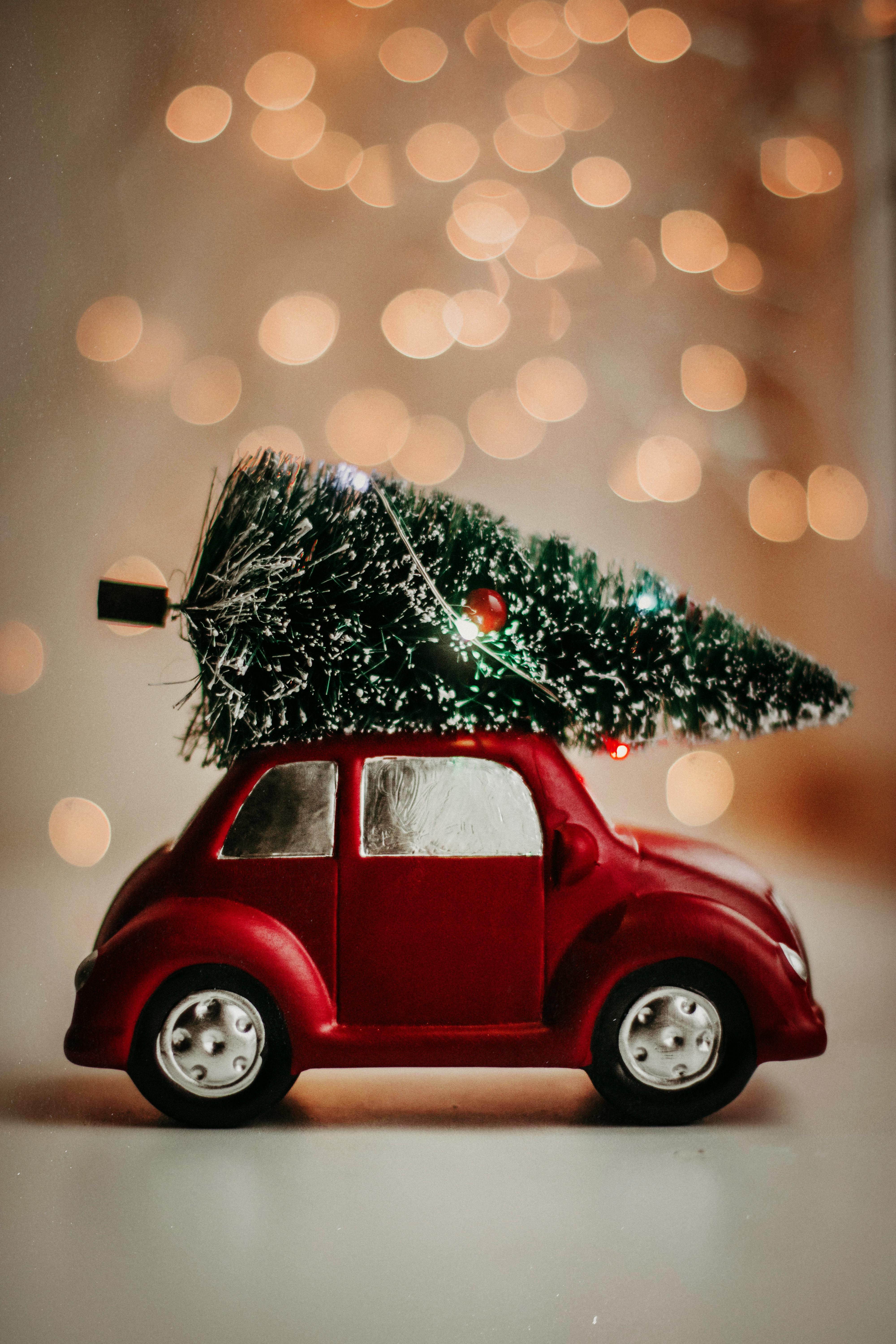 A red toy car is carrying a small Christmas tree, the background is warm-toned lights. - wallpaper image