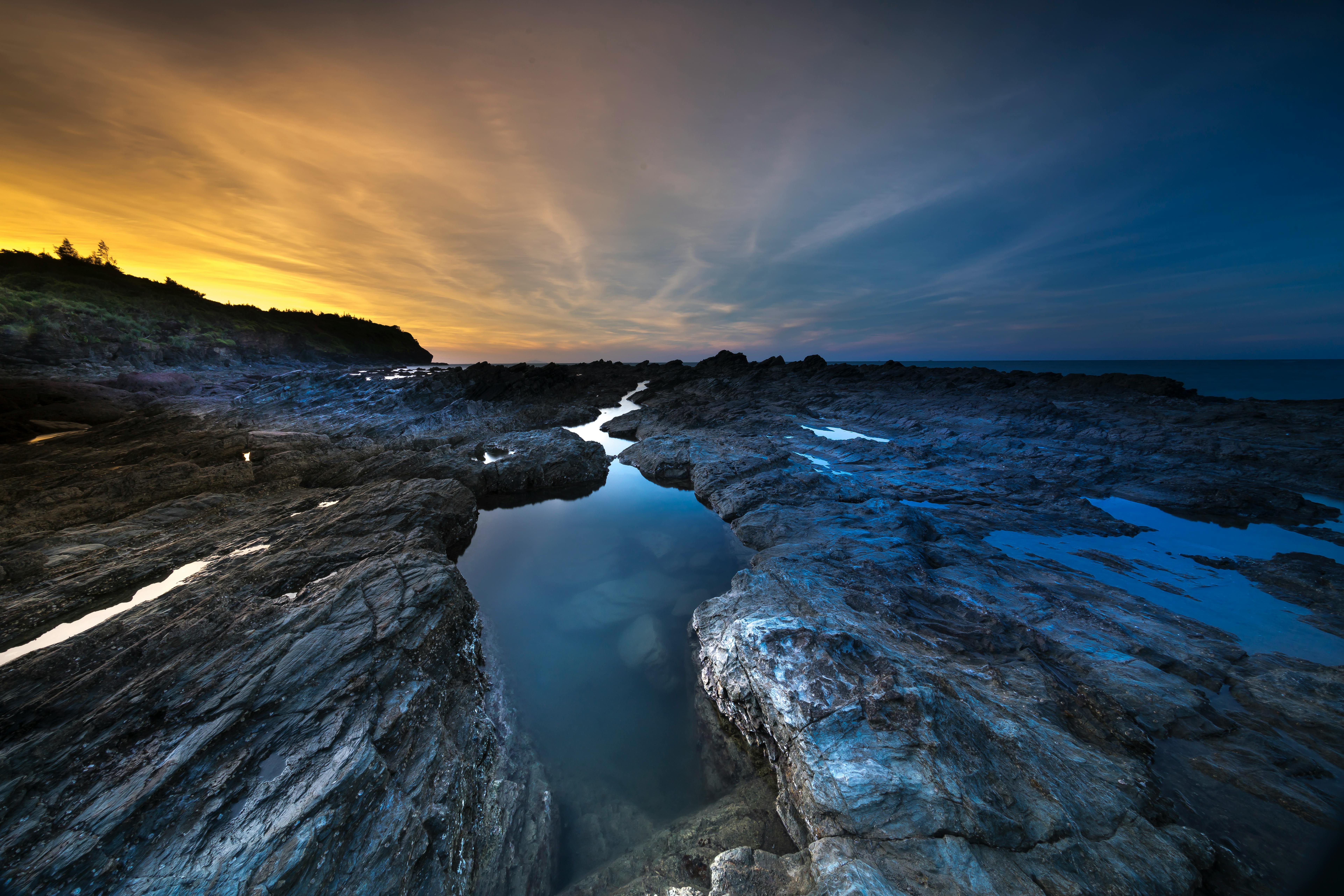 The sunset casts a gradient of orange and blue across the sky, reflecting in the tide pools formed along the rocky coast. - free wallpaper image