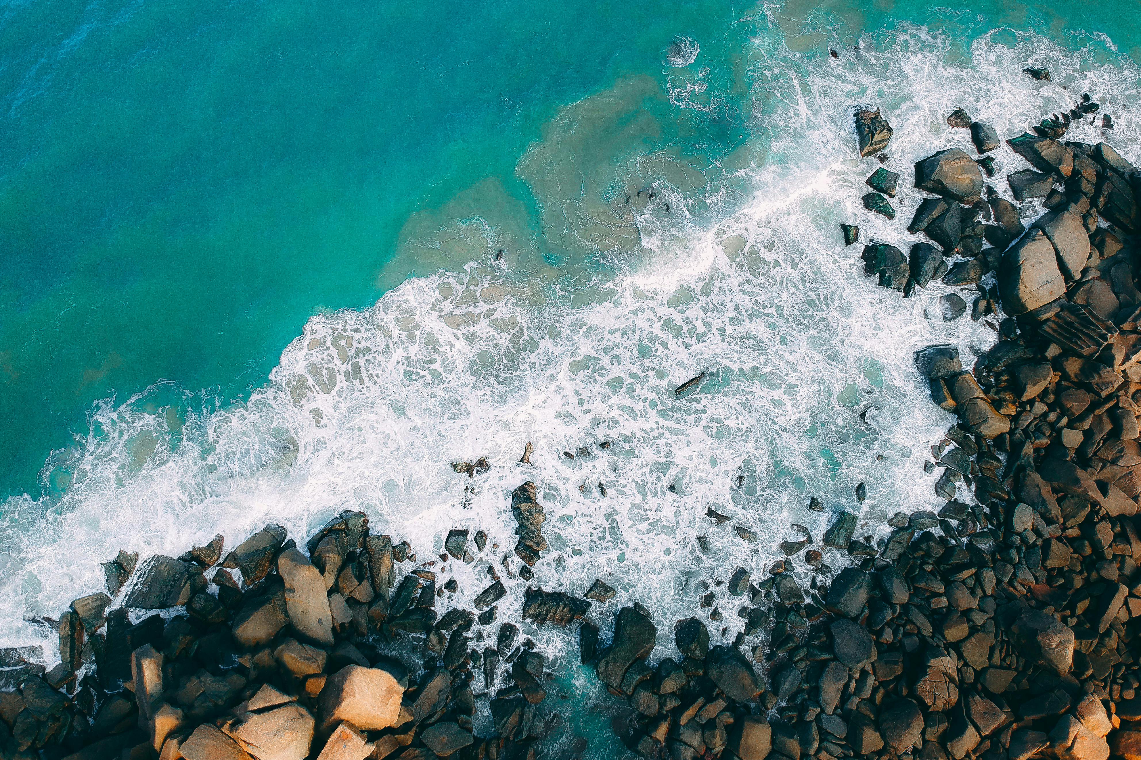 An aerial view of seaside rocks, the waves crashing against the rocks, forming white foam. - free wallpaper image