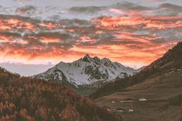 Snow-capped mountains with a warm orange sunset sky, there are some buildings on the hillside in the distance. - free wallpaper image