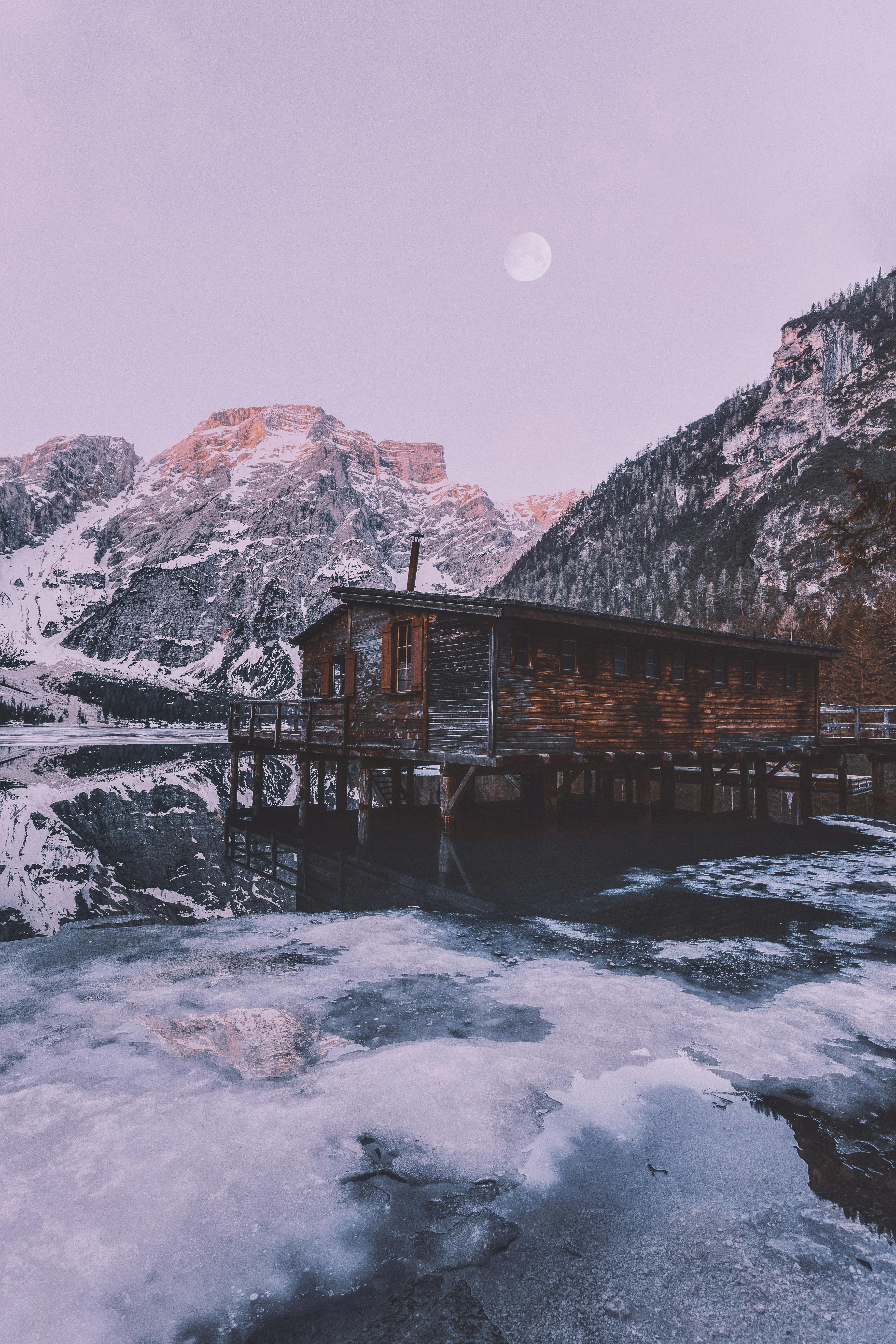 A wooden cabin sits on the edge of a lake surrounded by snow-capped mountains, the lake is frozen over and there is a full moon in the sky. - free wallpaper image