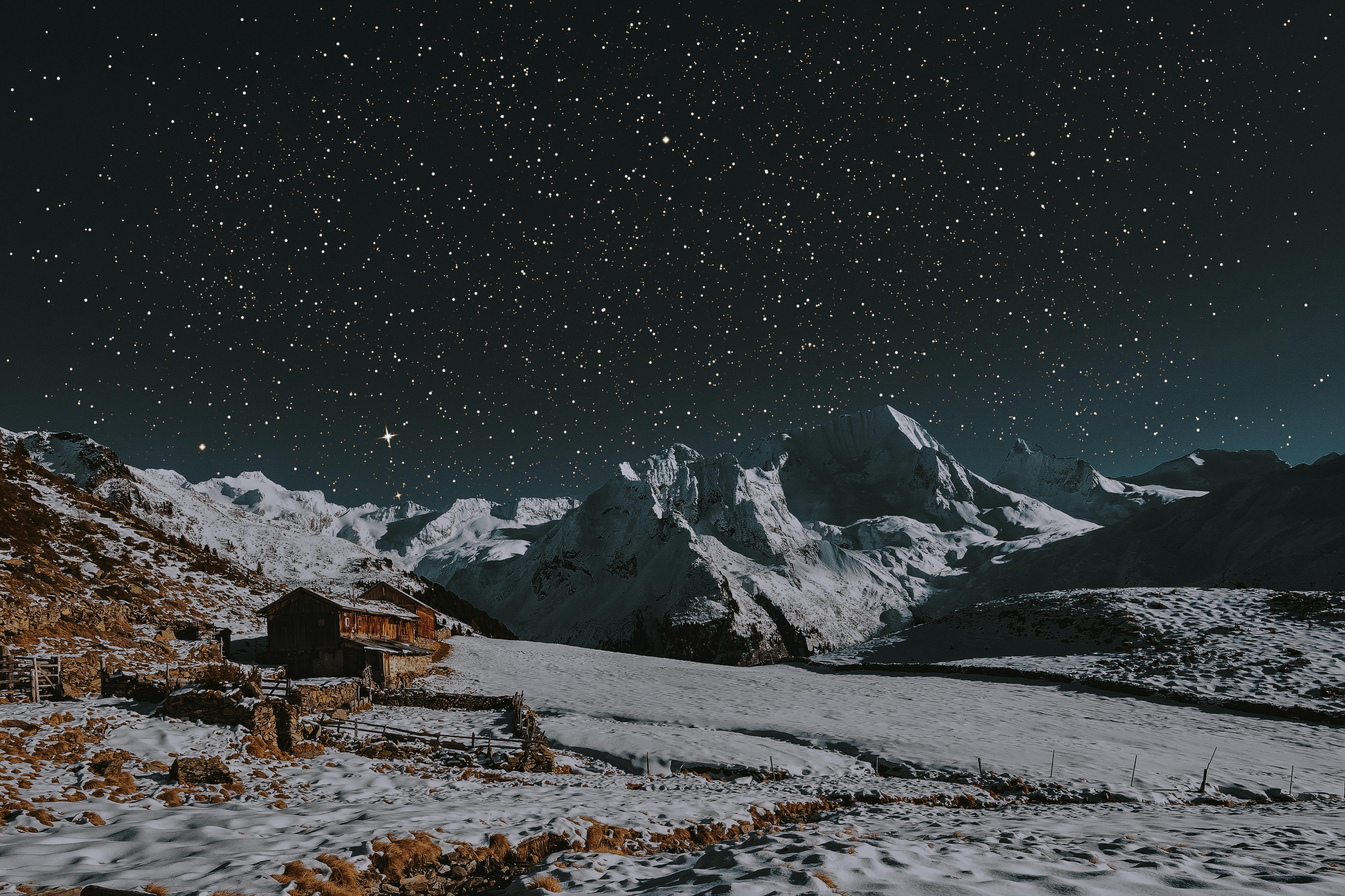 At night, under a starry sky, a snow-capped mountain stands tall, a wooden house stands at the foot of the mountain, surrounded by snow-covered land. - free wallpaper image