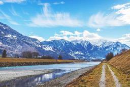 The snow-capped mountains in the distance stretch endlessly, at the foot of the mountains is a river, a small path runs along the river, the sky is blue, the clouds drift by. - free wallpaper image