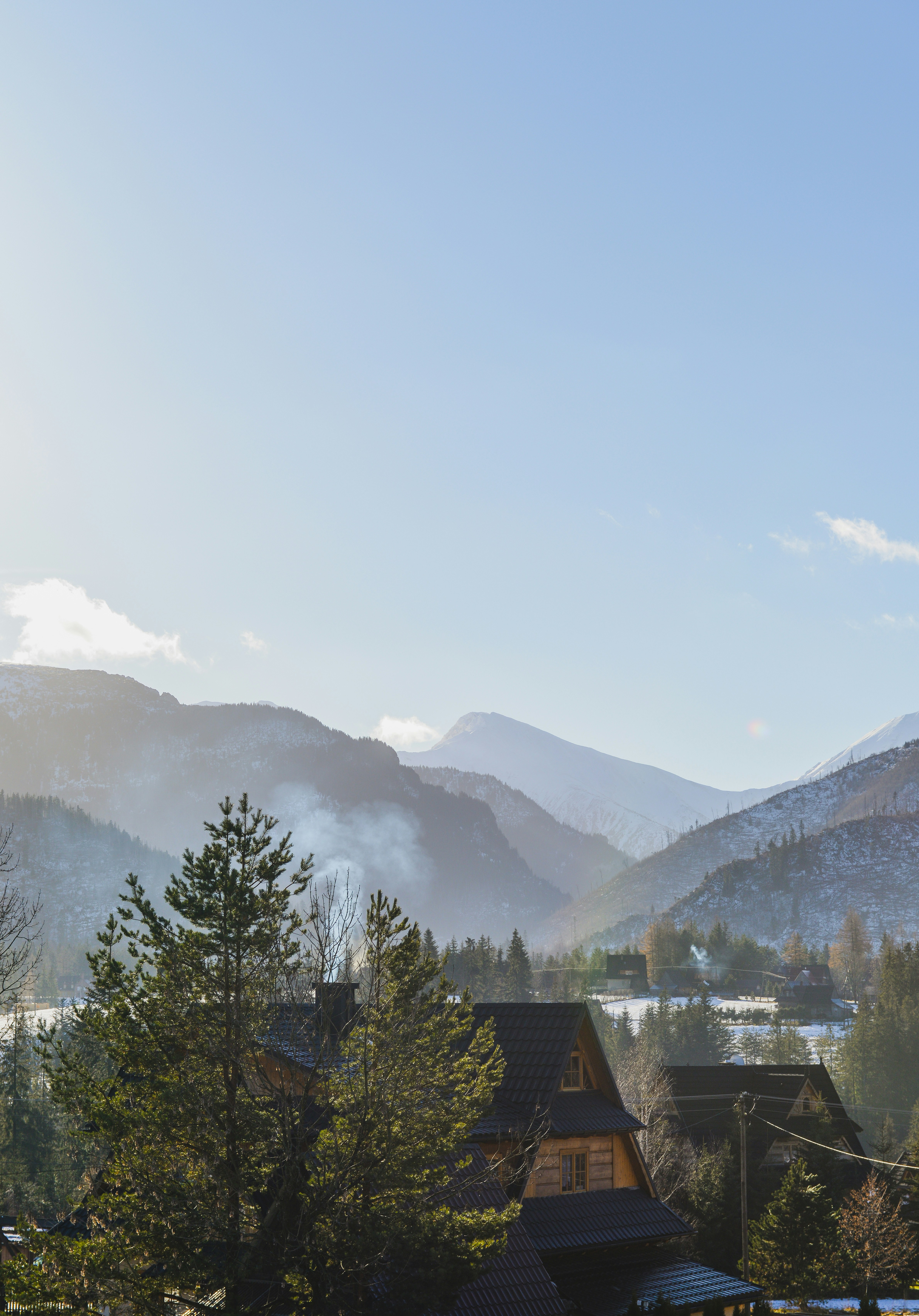 A mountain range surrounds the valley, at the foot of the mountain there is a forest and a few wooden houses, smoke rising from the roofs, the sky is clear and the sun is shining. - free wallpaper image