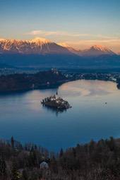 A lake with an island and a church on it, surrounded by mountains with snow on top. - free wallpaper image