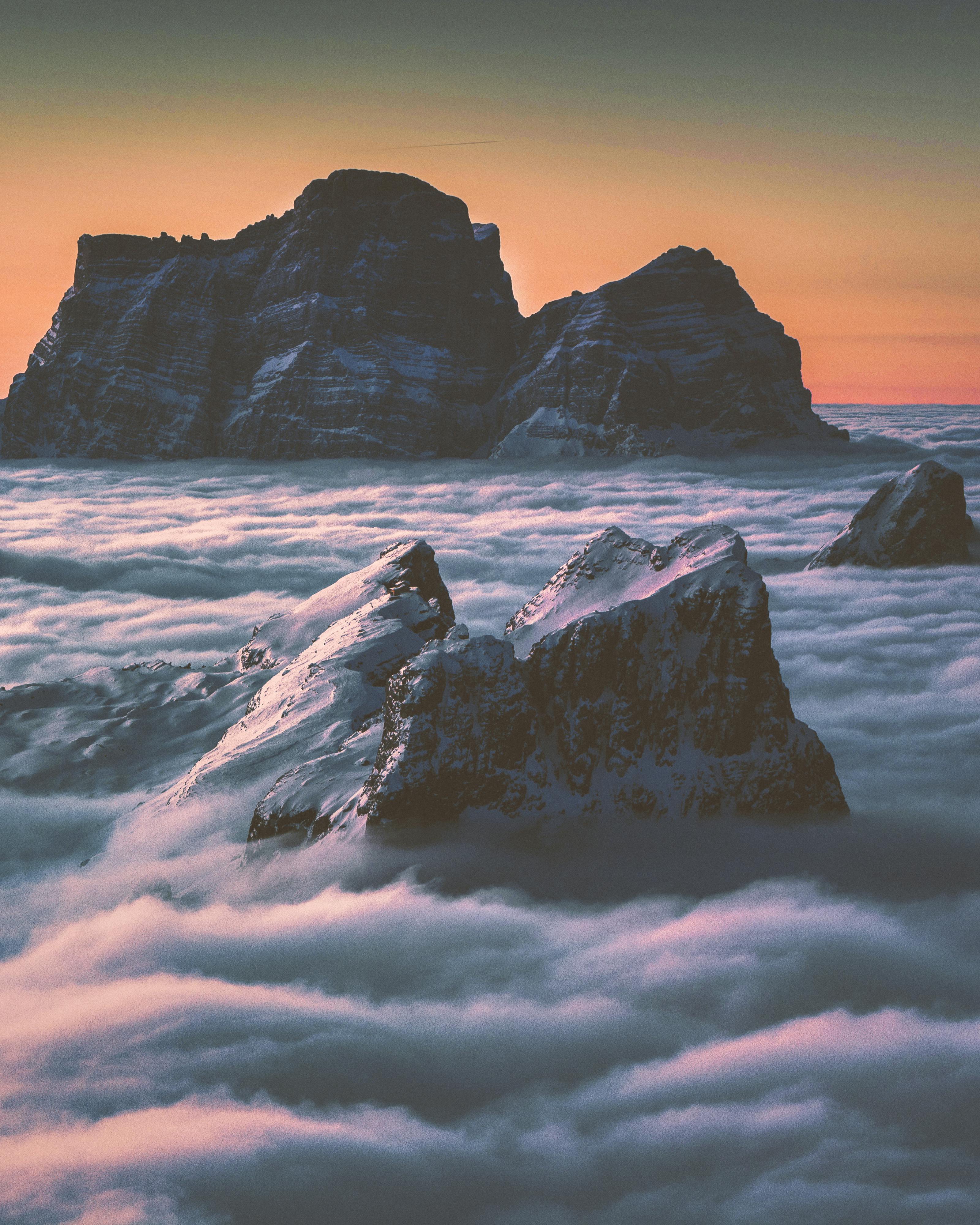 Several mountain peaks stand tall above the clouds, the setting sun paints the sky with its glow. - free wallpaper image