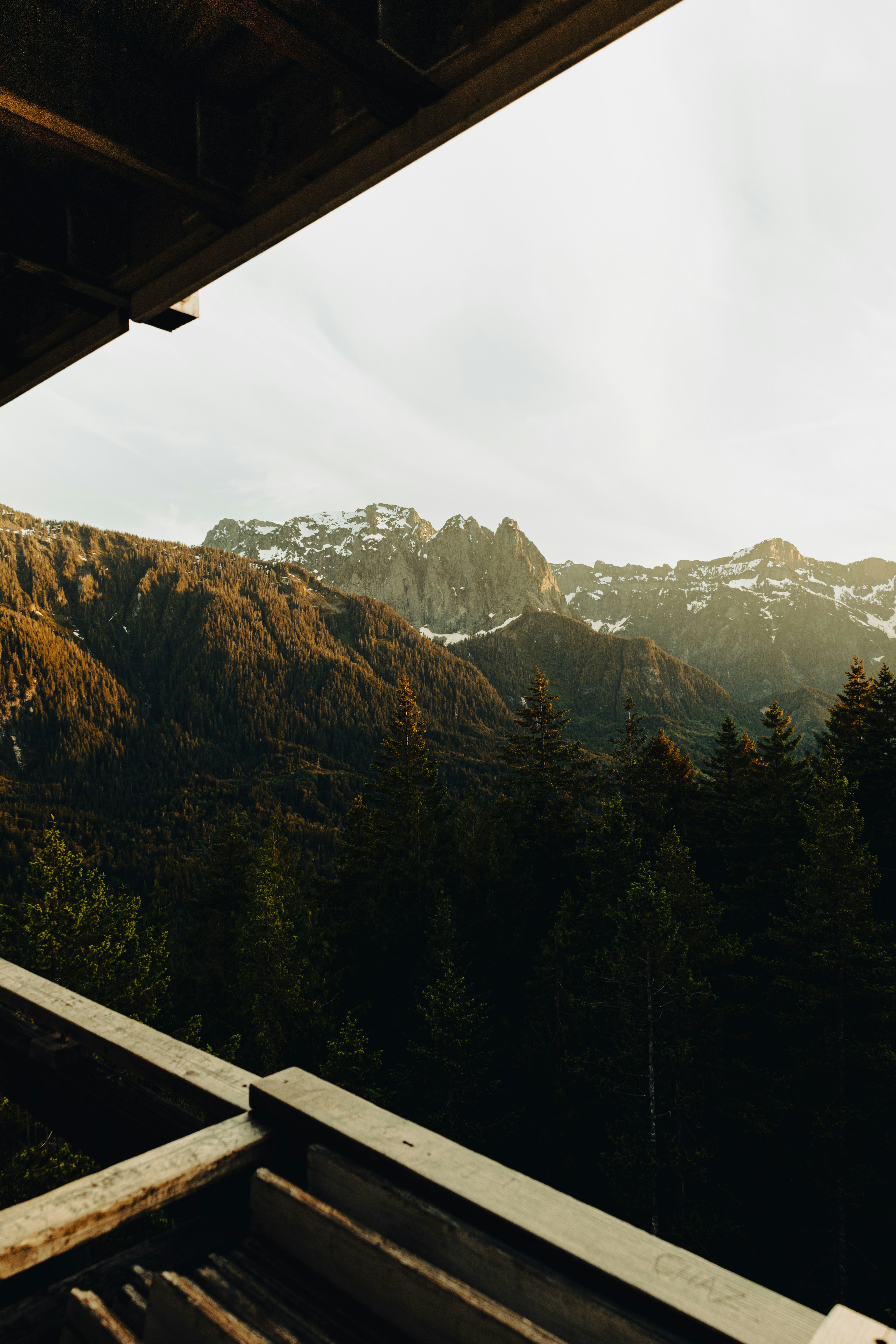 A view of snow-capped mountains from a wooden balcony, with lush forests covering the mountainsides, a bright blue sky, and a breathtaking view. - free wallpaper image