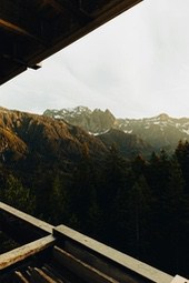 A view of snow-capped mountains from a wooden balcony, with lush forests covering the mountainsides, a bright blue sky, and a breathtaking view. - free wallpaper image