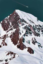 A snowy mountain peak with exposed rock, an airplane flies over in the sky. - free wallpaper image