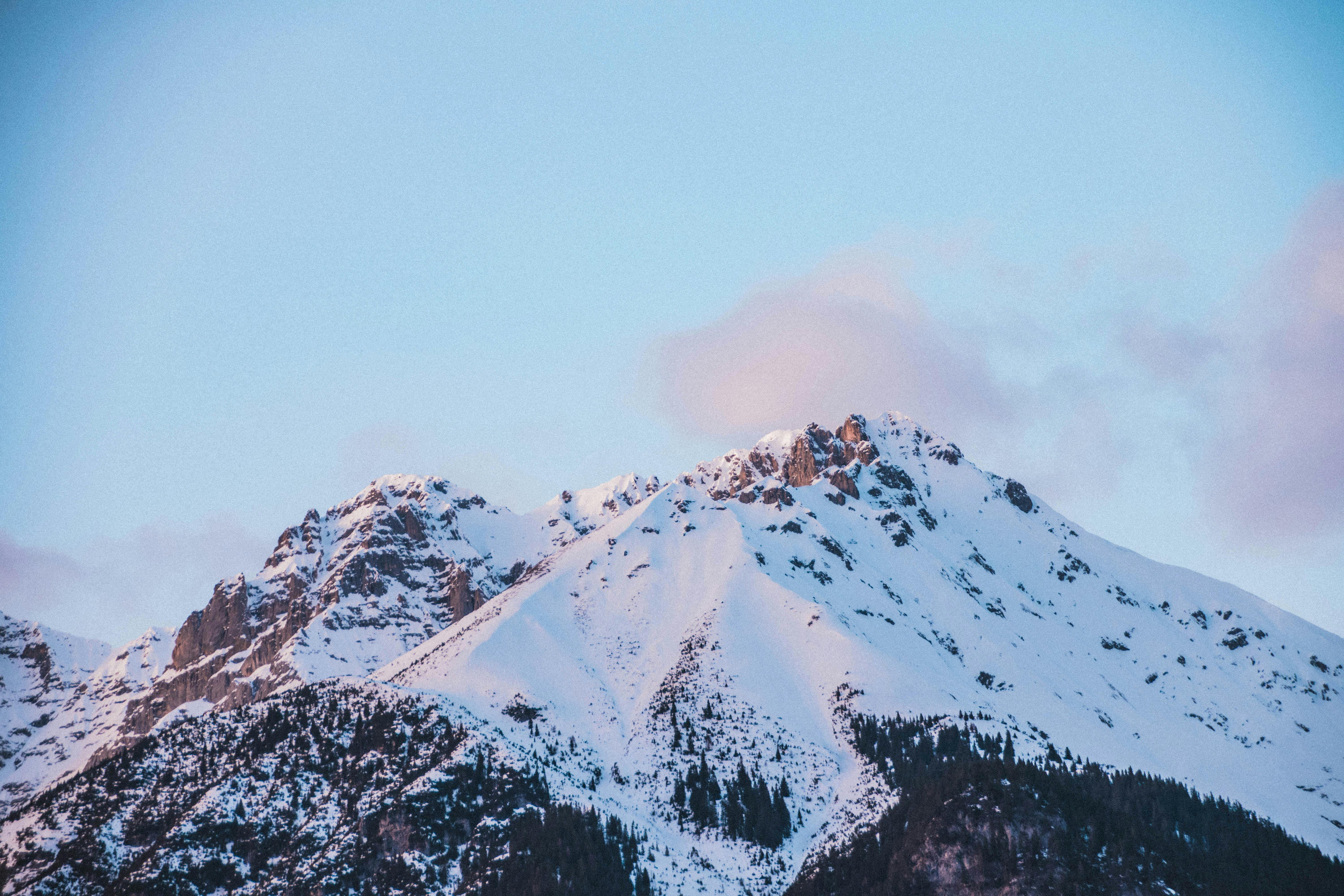 The top of the snowy mountain is covered with white snow, the mountain slopes are covered with trees, the sky is light blue and the clouds are sparse. - free wallpaper image