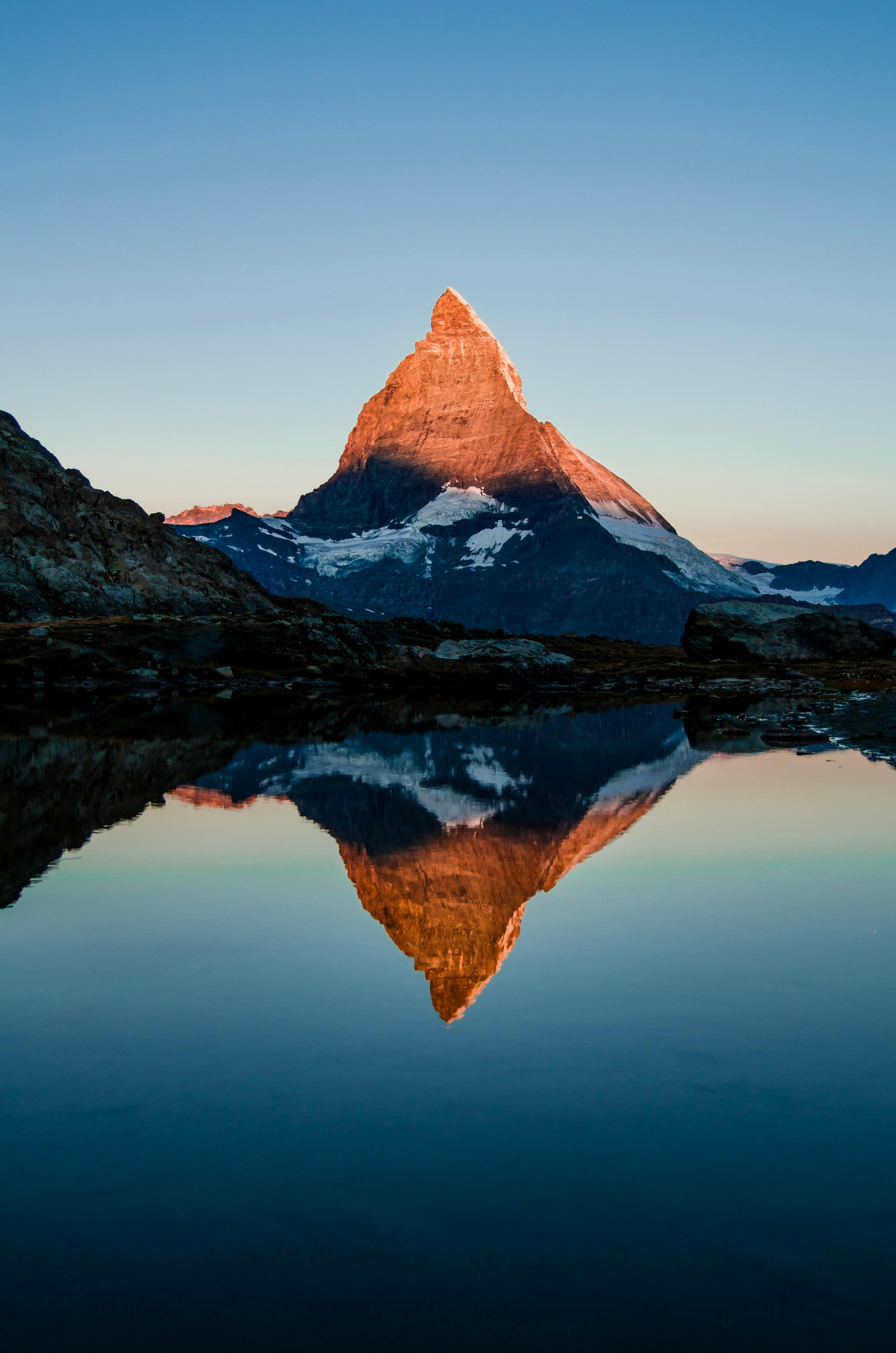 A mountain peak is reflected in a still lake. The sky is blue, the mountain is brown and the top of the mountain is covered with white snow. - free wallpaper image