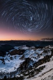 The mountaintop is covered with snow, the distant mountains are a faint red in the afterglow of the sunset, and the night sky is dotted with stars, forming a spiral star trail. - free wallpaper image