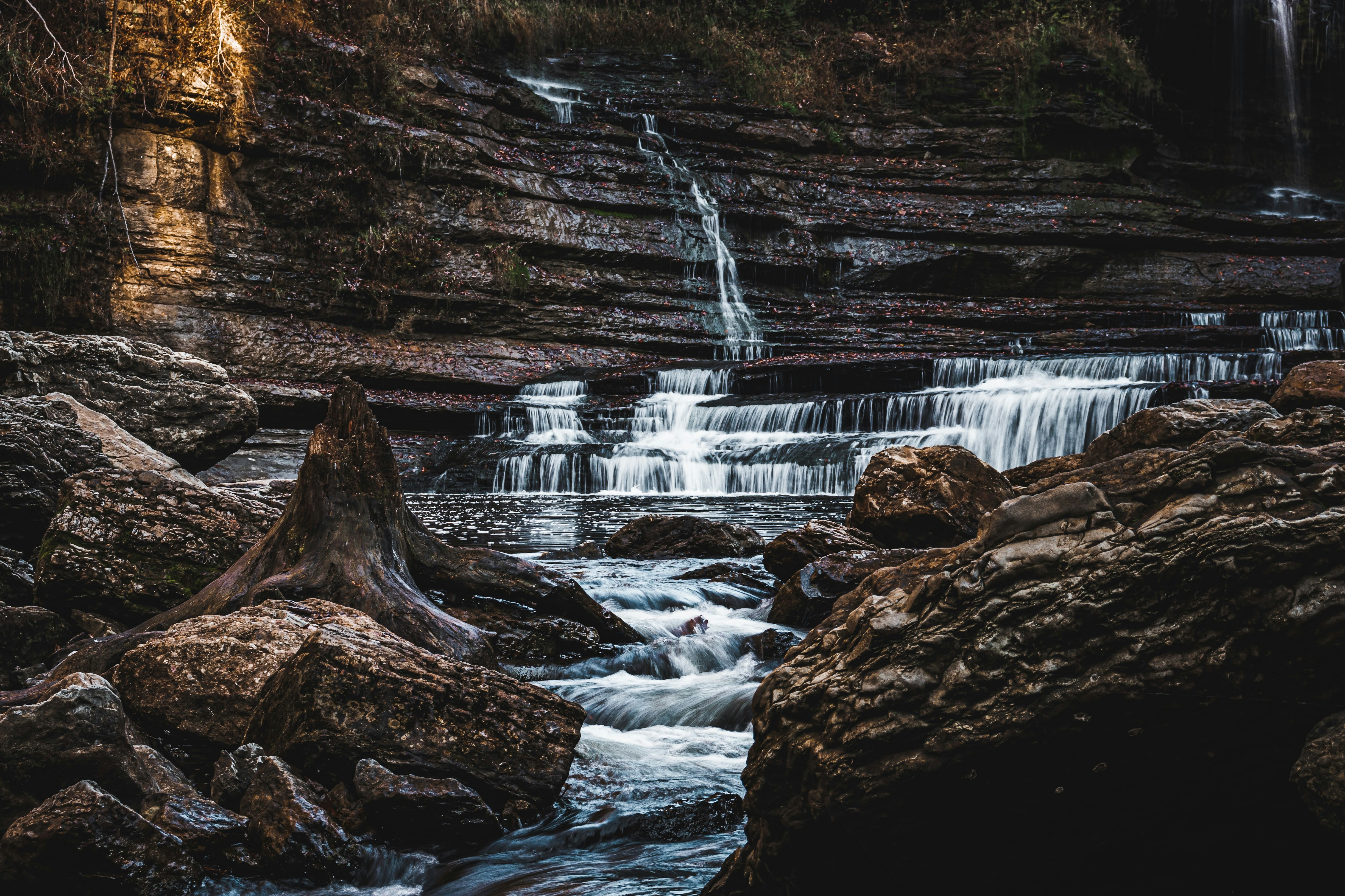 A stream flows through rocks, forming a cascade of small waterfalls, sunlight shines on the rocks, creating warm tones of light and shadow. - free wallpaper image