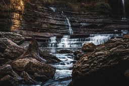 A stream flows through rocks, forming a cascade of small waterfalls, sunlight shines on the rocks, creating warm tones of light and shadow. - free wallpaper image